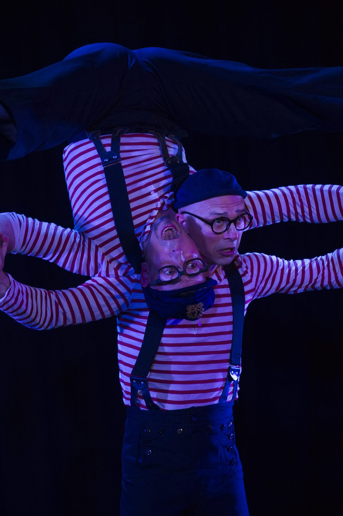 One performer held inverted across the other's shoulders, arms interlocked, in deep blue light